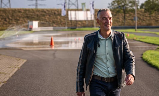 Man van ongeveer 60 jaar met zwarte leren jas loopt zelfverzekerd over het oefenterrein van de rijschool.