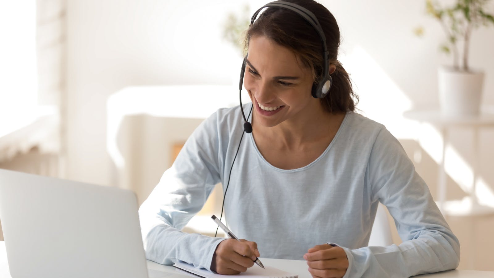 een vrouw zit met headset op achter een laptop en maakt aantekeningen op papier