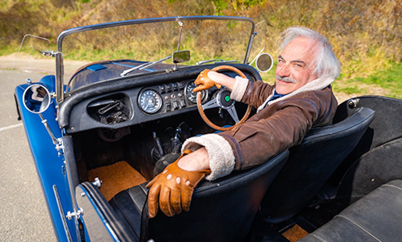 Claude zit, met zijn arm nonchalant over de bijrijdersstoel, in zijn Morgan