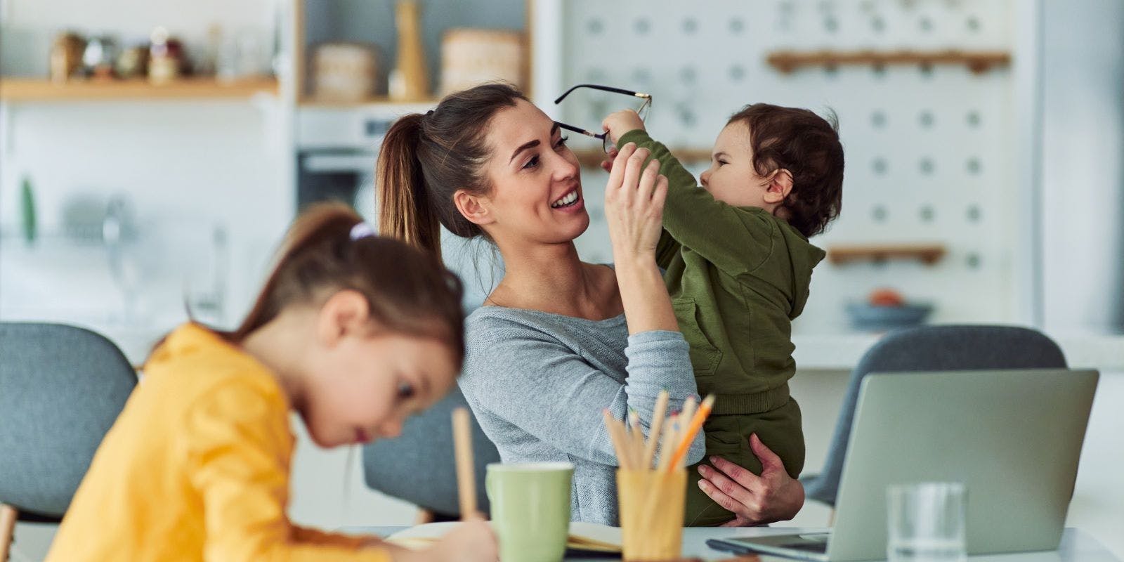Vrouw zit aan tafel en zoekt op de laptop hoe het zit met een kapotte bril en de verzekering. 1 van haar 2 kinderen grijpt naar haar bril.