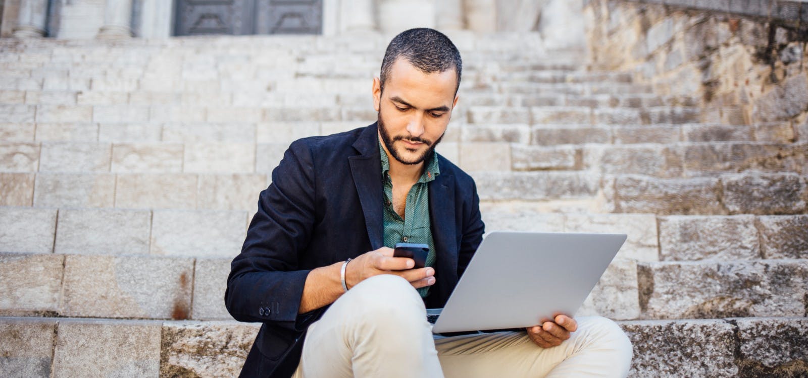 Man zit op stenen trap met zijn laptop en kijkt op zijn mobiele telefoon