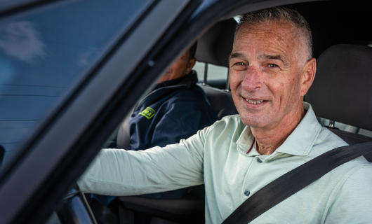 Man van ongeveer 60 jaar zit in lichtgroene blouse achter het stuur en adviseert de lezer hoe je een autoverzekering afsluit als beginnend bestuurder