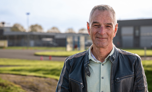 Man van ongeveer 60 jaar staat rechts in beeld lacht richting de camera