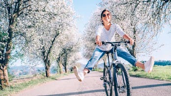 een vrouw slingert haar benen vrolijk vooruit op de fiets, op de achtergrond staan bomen vol in de bloesems