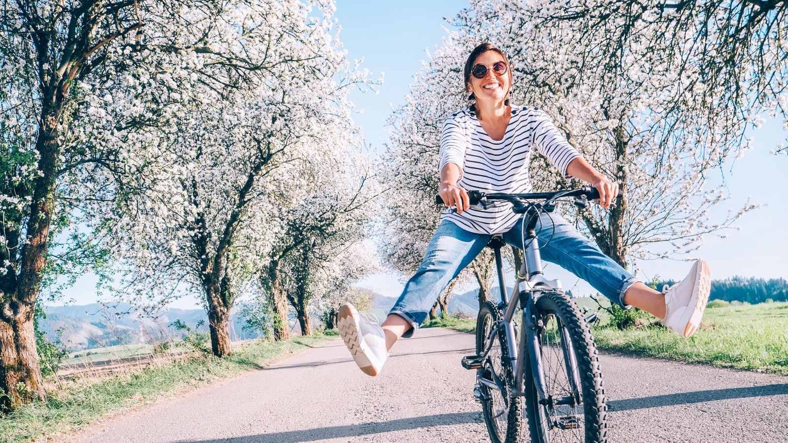 een vrouw slingert haar benen vrolijk vooruit op de fiets, op de achtergrond staan bomen vol in de bloesems