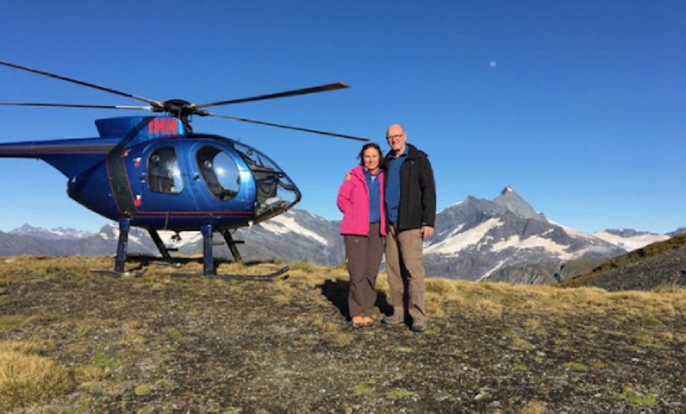 een echtpaar staat met een helicopter op een helling, met op de achtergrond bergtoppen
