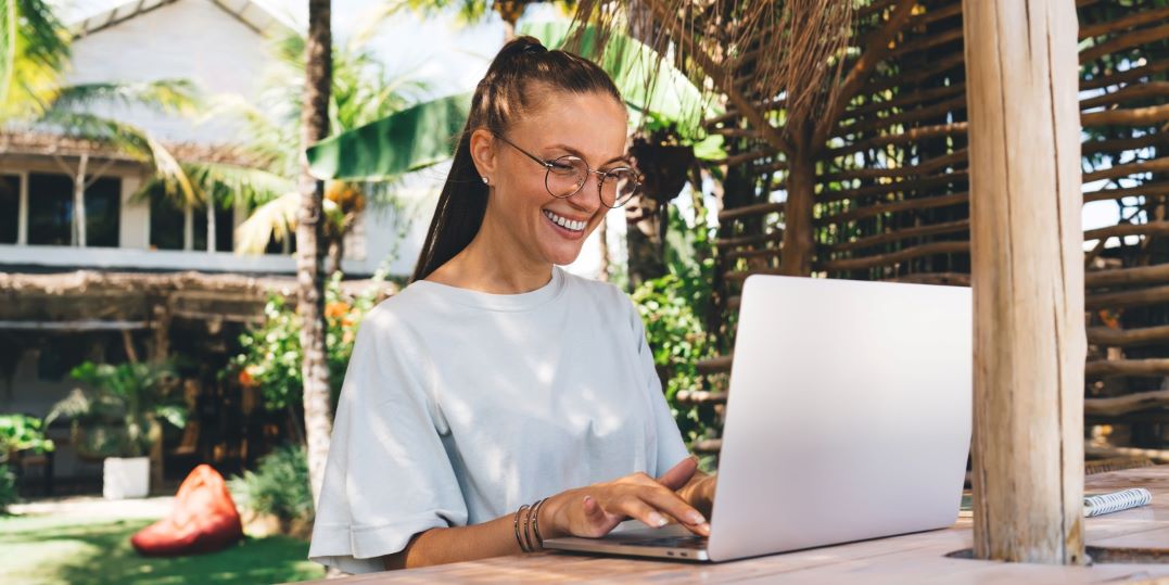 Vrouw (31 jaar) met bril en wit shirt zit aan houten tafel, buiten in een zonnige tuin. Ze vertelt via Skype hoe je digital nomad kan worden.