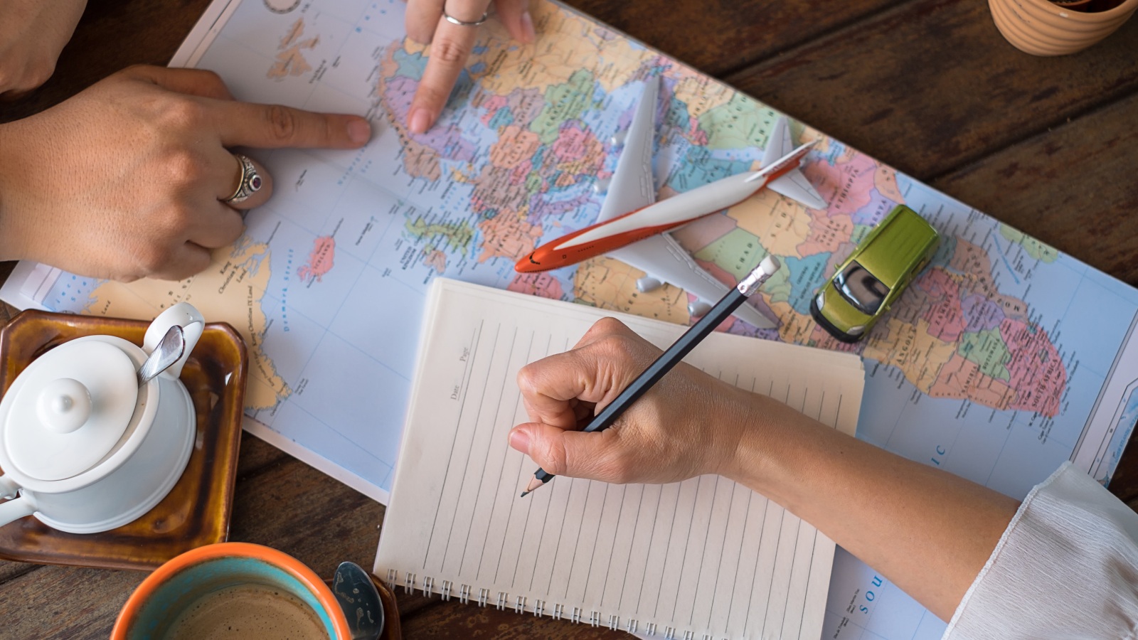 er wordt een reis gepland; op de tafel ligt een vliegtuigje, een autootje, iemand noteert in een schrijfblok en anderen wijzen op een wereldkaart.