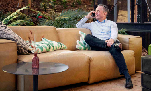 Man in blauwe blouse zit op bank met telefoon in hand en vertelt dat een goede reisverzekering ontzorgt tijdens je studie