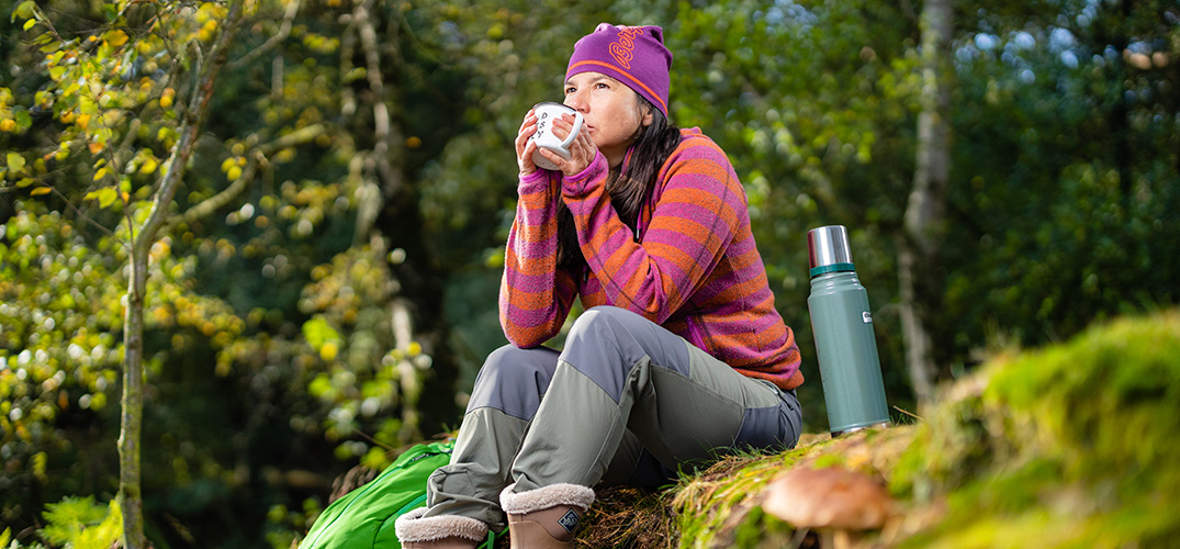 Susan neemt zittend in het bos slokjes uit een beker, met op de voorgrond een thermosfles