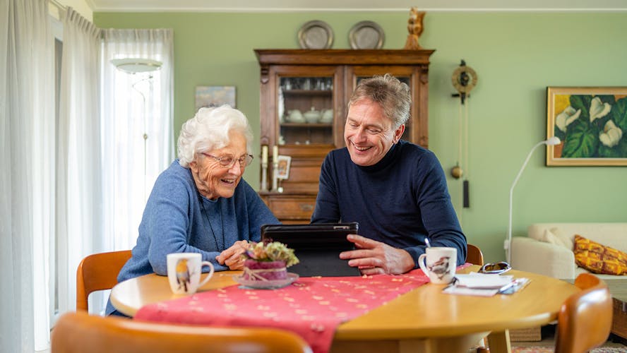 Gerben zit met zijn schoonmoeder aan de eettafel, beiden lachen om wat ze zien op een tablet