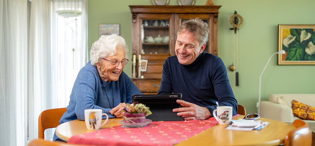 Gerben en zijn schoonmoeder zitten aan de eettafel en kijken lachend naar het scherm van een tablet