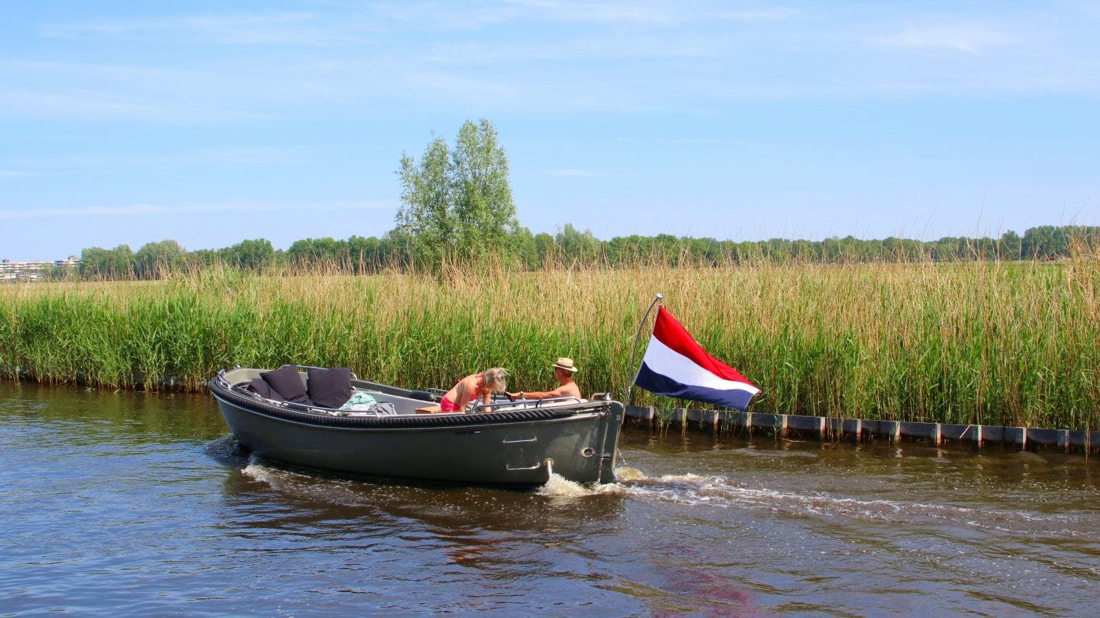 twee personen varen in een sloepje met de Nederlandse vlag door een zonnig groen landschap