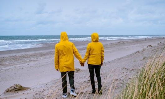 twee personen in gele regenjassen lopen hand in hand door een bewolkt duinlandschap