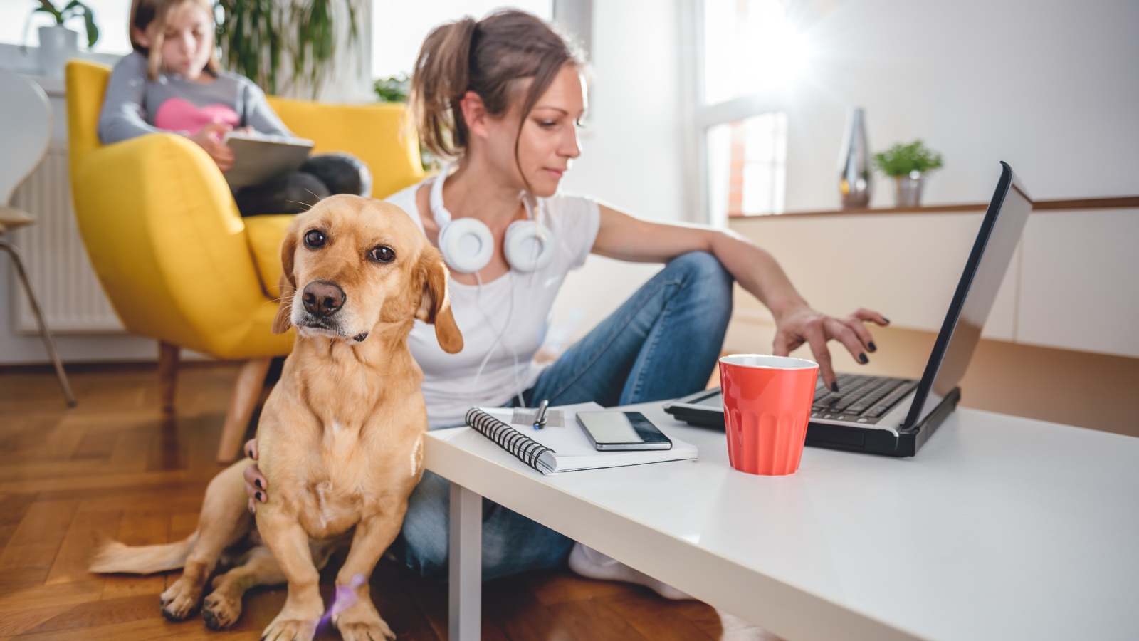 Vrouw werkt huis op de laptop, in de woonkamer aan de salontafel, met hond en kind in de buurt.