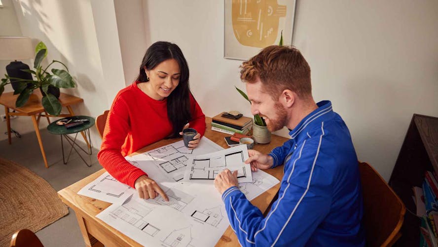 Een man en een vrouw zitten aan tafel en bekijken samen uitgeprinte plattegronden.