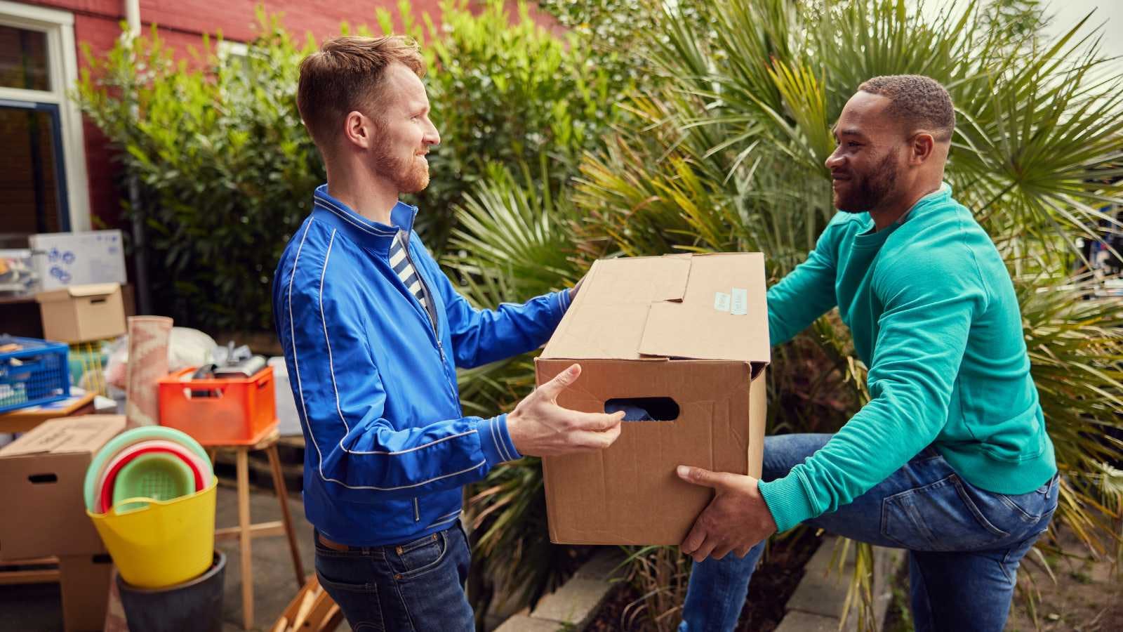 Een man helpt zijn vriend en neemt de verhuisdoos van hem over. Op de achtergrond staan nog meer verhuisspullen.