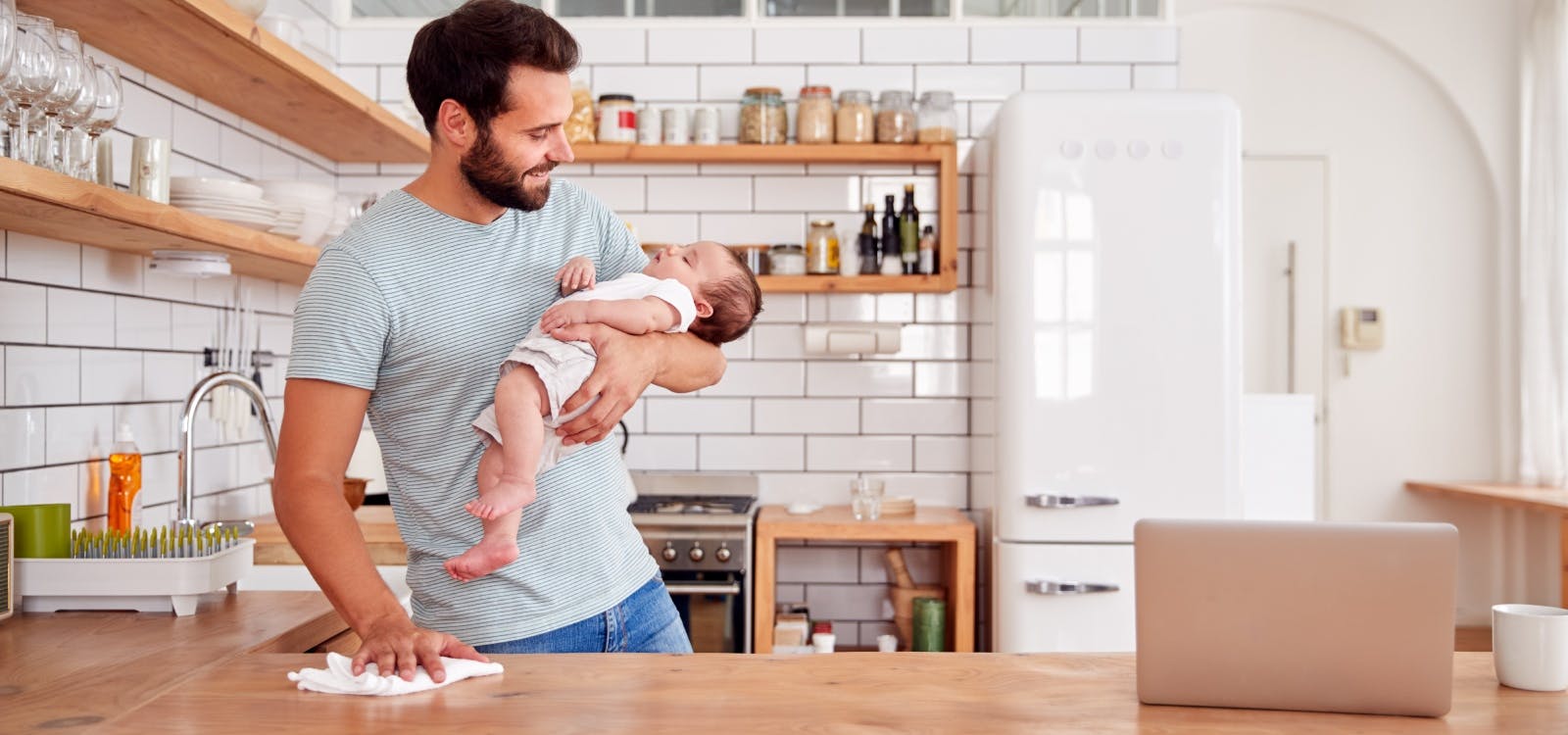 een vader lacht tegen de baby in zijn ene arm, terwijl hij met de andere hand het aanrecht schoonmaakt