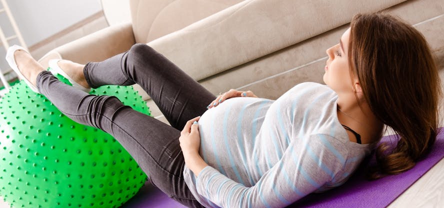 een zwangere vrouw ligt op een paarse yoga-mat met haar benen op een groene gymbal