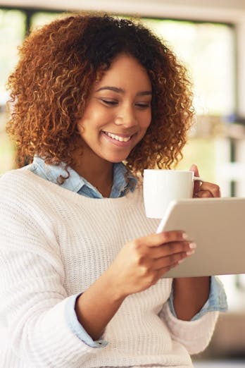 Blije vrouw met bruin haar en witte trui drinkt koffie en bekijkt op haar tablet de Jaarcijfers zorg 2023 