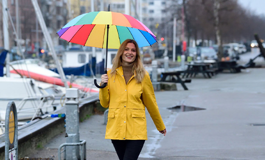 Blonde vrouw van 26 jaar loopt in felgele regenjas en regenboog gekleurde paraplu door de regen langs de kade met aangemeerde boten. 