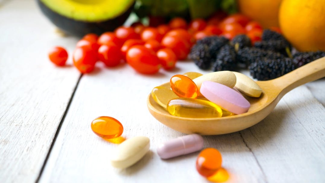 een lepel vol verschillende vormen en kleuren pillen, met op de achtergrond verschillende soorten fruit