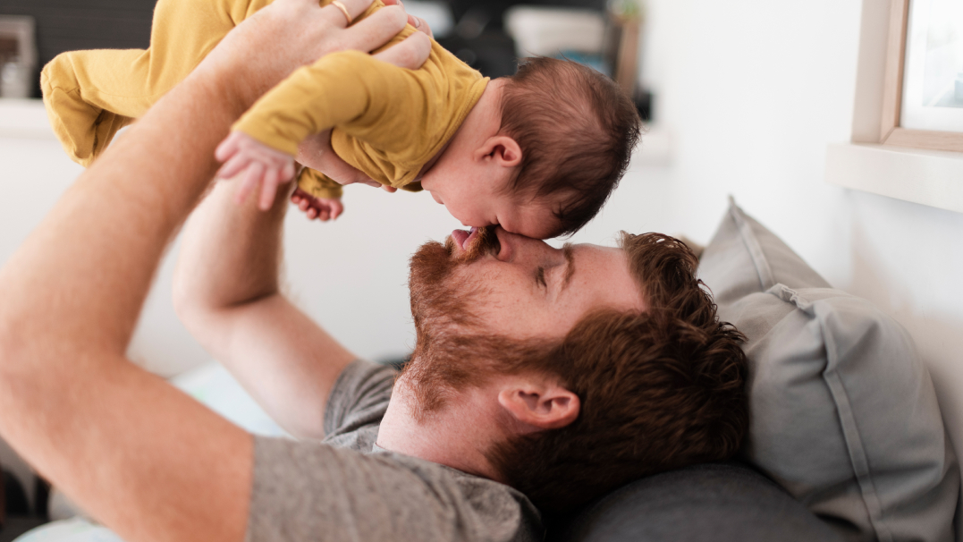 Een kersverse vader zit op de bank en houdt zijn baby speels boven zich in de lucht voor een kus.