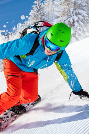 Snowboarder met groene muts, blauwe jas en rode broek, op een sneeuwwitte helling en strakblauwe lucht