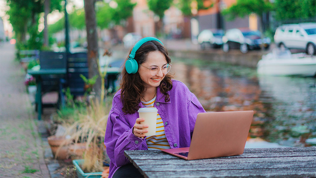 Een vrouw met vrolijk gekleurde kleding zit aan de gracht. Ze draagt een bril en een grote glimlach. Met een kop koffie in de hand is ze aan het werk op haar laptop. 