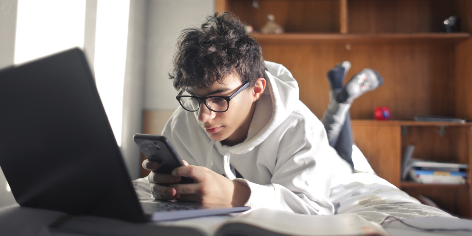 Student met bril en grijze sweater ligt ontspannen op bed tijdens het leren op zijn laptop. Hij vraagt ondertussen zorgtoeslag aan op zijn telefoon.